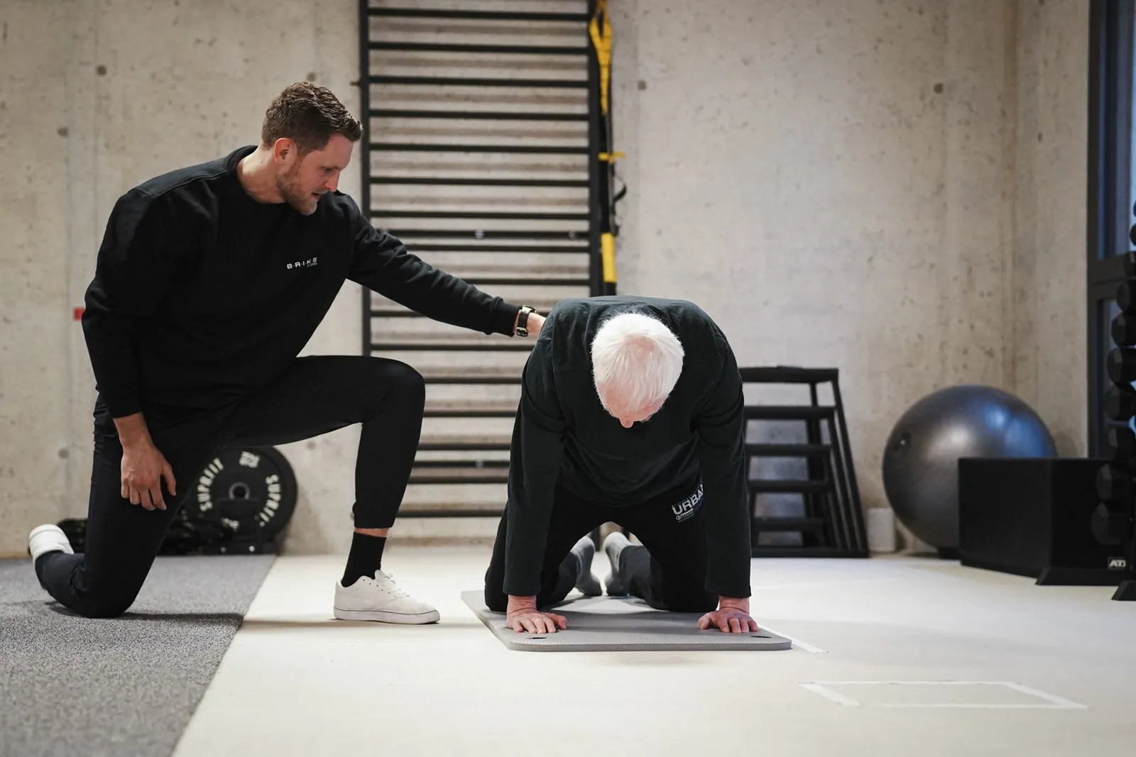 Trainer begleitet einen Senior bei einer Übung im Vierfüßlerstand auf der Matte im BRIKS Studio